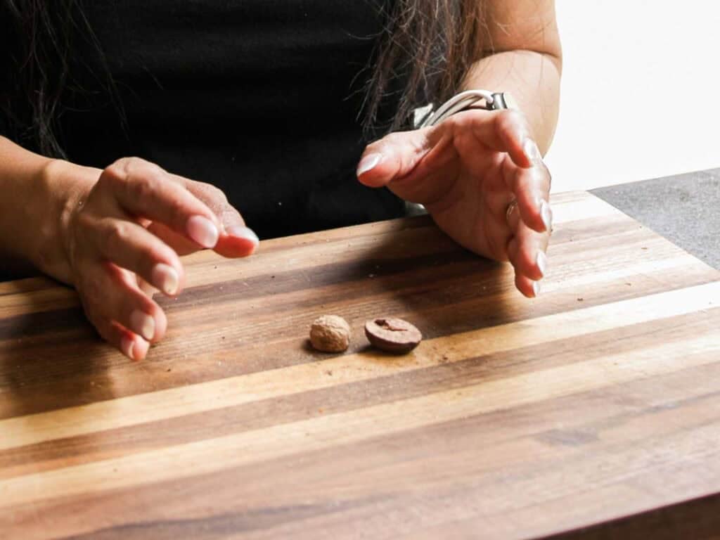A person with long hair gestures over a striped wooden cutting board with two whole nutmegs, preparing ingredients for a vegan bolognese. Only their hands and part of their torso are visible.